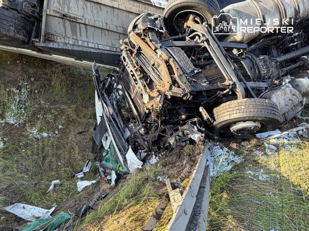 W przewróconym pojeździe dostawczym widoczne są uszkodzone elementy silnika oraz zniszczona karoseria, otoczone trawą.