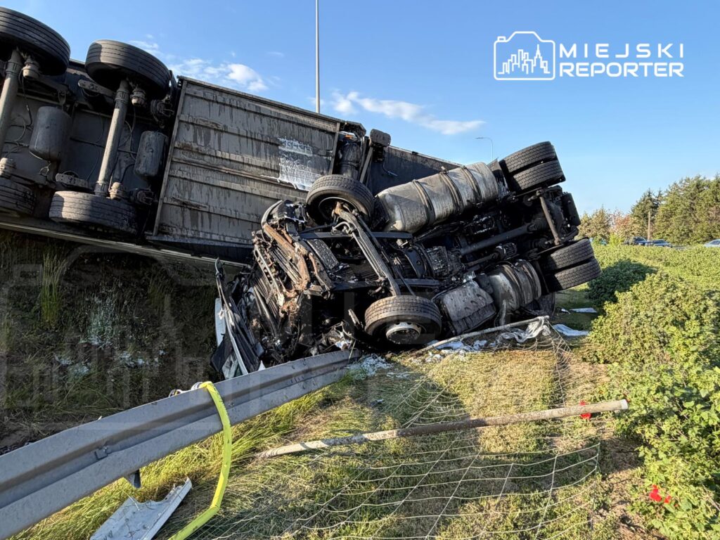 Przewrócona ciężarówka leży na boku wzdłuż drogi, obok metalowego barierek, w otoczeniu zieleni i niebieskiego nieba.