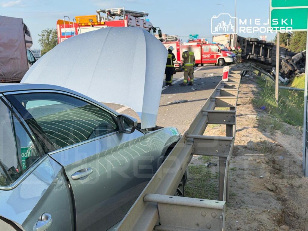 Samochód z otwartą maską stoi na poboczu drogi, w tle widać służby ratunkowe i przewrócony pojazd.