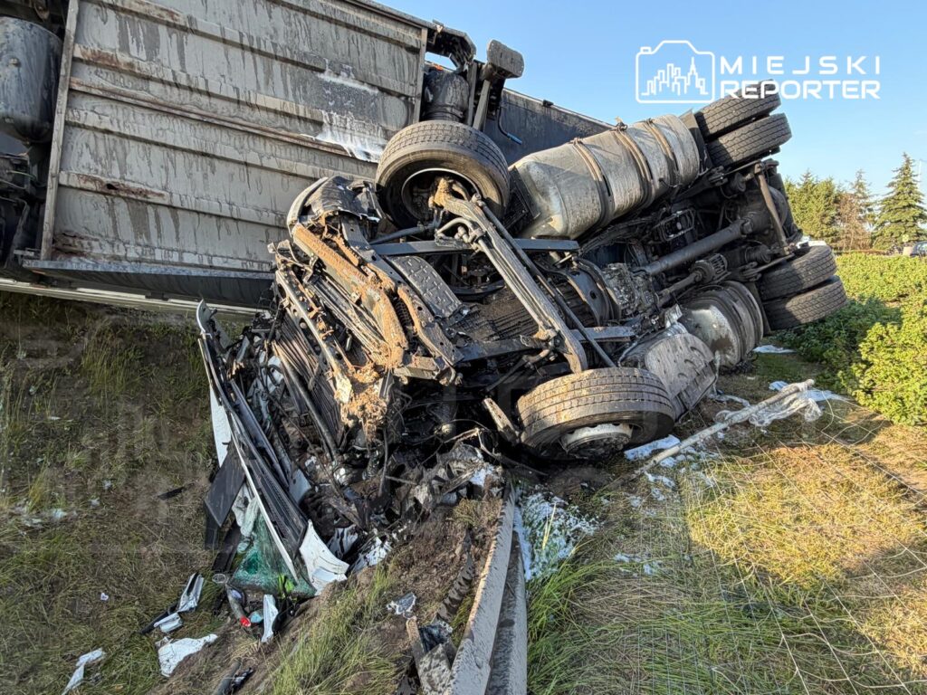 Wielka ciężarówka leżąca na boku w rowie, z uszkodzoną kabiną i porozrzucanymi fragmentami pojazdu.