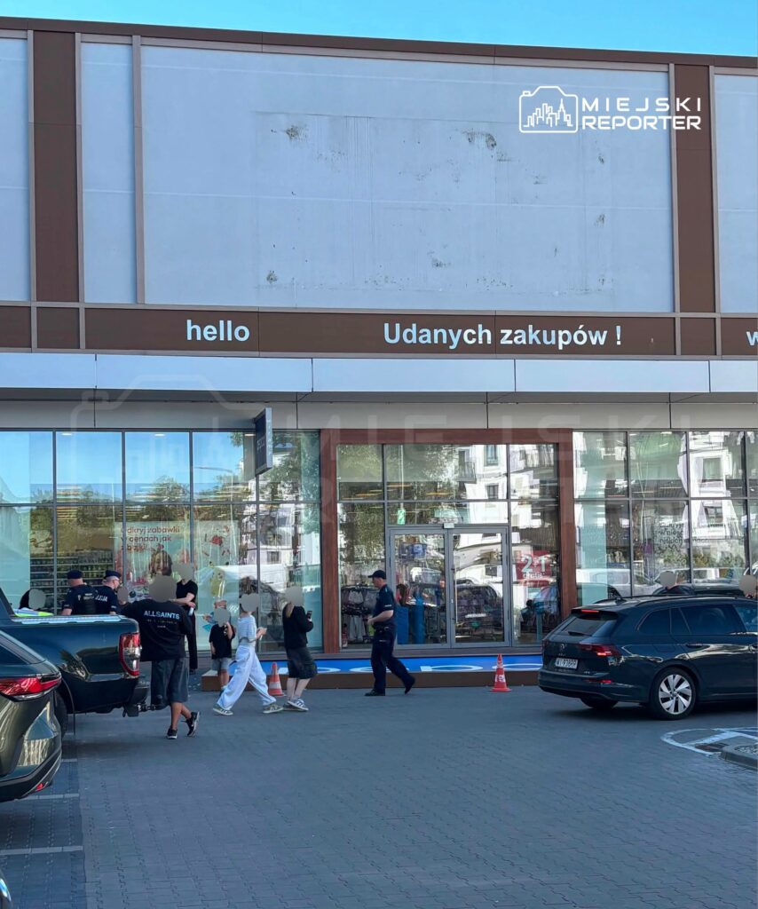Grupa osób z torbami przechodzi obok sklepu, a policjanci prowadzą rozmowy na parkingu przed budynkiem.