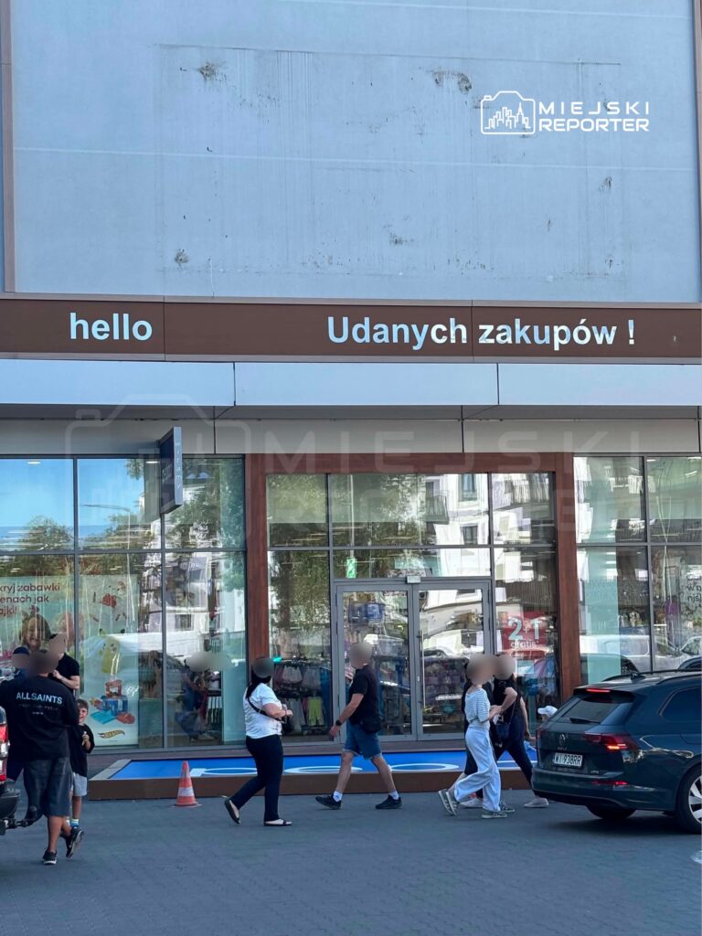 Grupa osób spaceruje przed nowoczesnym budynkiem handlowym z dużymi witrynami. Na elewacji widnieje napis: "hello" oraz "Udanych zakupów!".