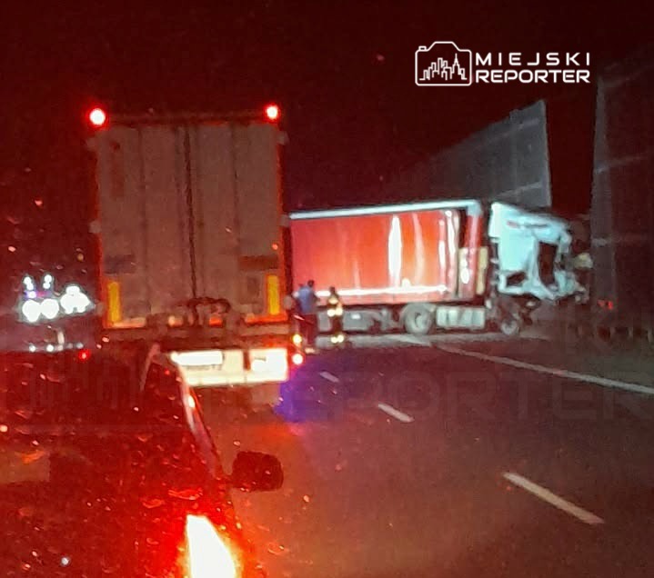 W nocy na drodze stoi uszkodzony biały ciężarówka, obok niej interweniują służby ratunkowe. W tle widać inne pojazdy.