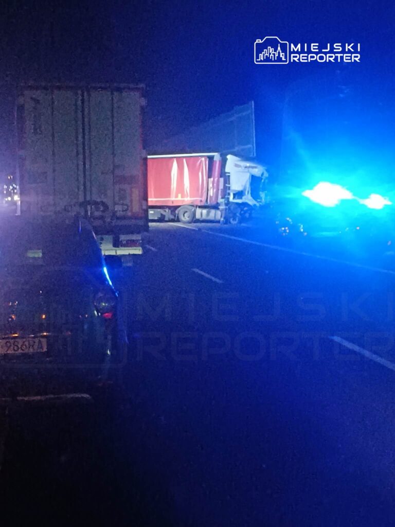 Nocna scena na drodze z wypadkiem, uszkodzony ciężarówka z czerwoną naczepą, niebieskie światła radiowozu.