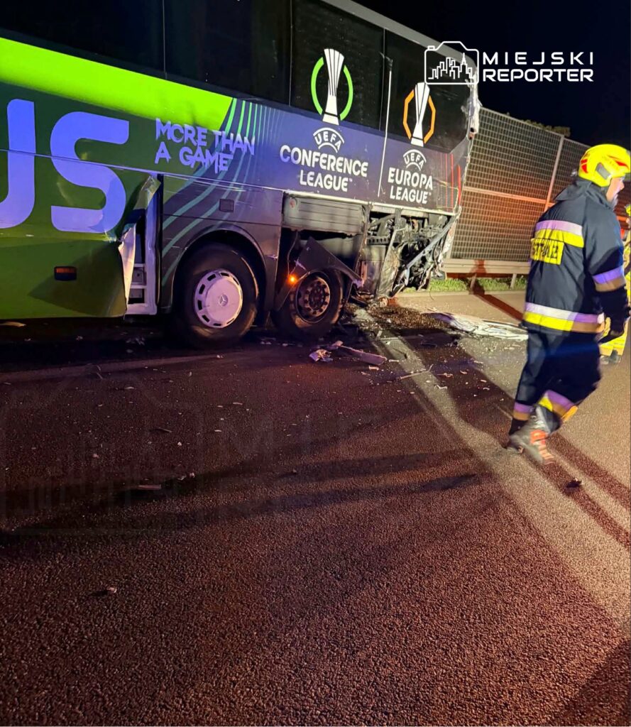 Autobus z oznaczeniami UEFA, uszkodzony po kolizji, stoi na drodze, a obok pracuje strażak w mundurze.