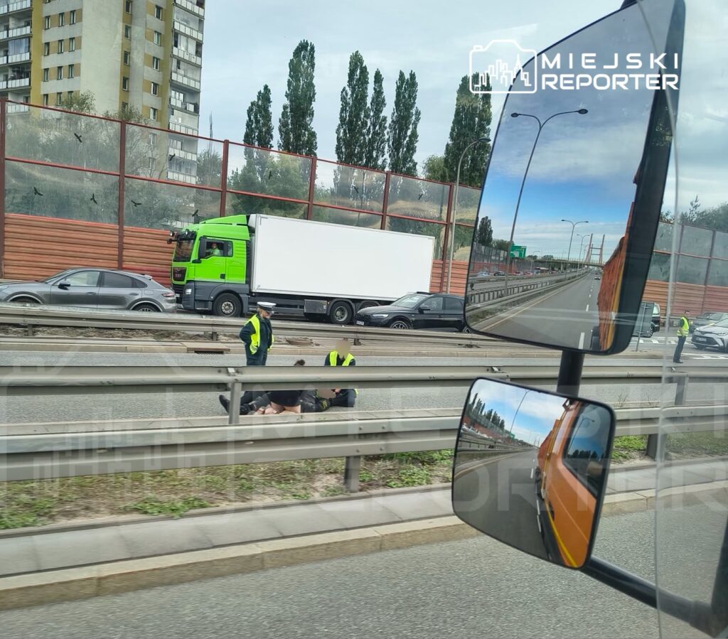 Na jezdni leżą dwie osoby, a obok nich pracują ratownicy w odblaskowych kamizelkach. W tle przejeżdżają pojazdy.