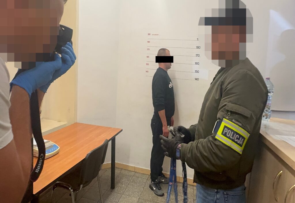 Mężczyzna w czarnym dresie stoi przy ścianie z podziałką wzrostu, a policjant w mundurze trzyma kajdanki i notuje informacje.
