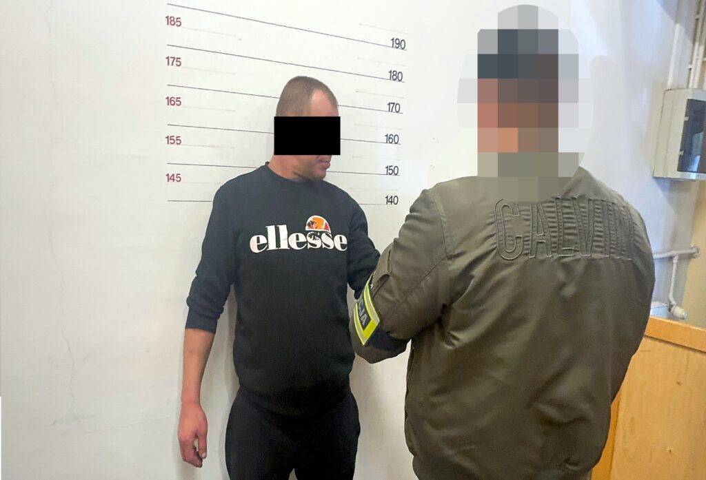 Mężczyzna w czarnej bluzie stoi przy ścianie z oznaczeniami wysokości, policjant z tyłu trzyma go za ramię.