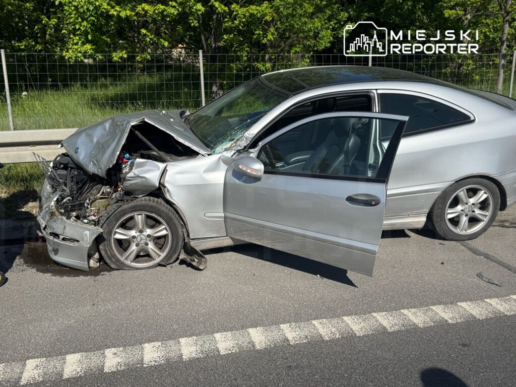 Zniszczony srebrny samochód z wgniecionym przodem stoi na drodze, a drzwi są otwarte.