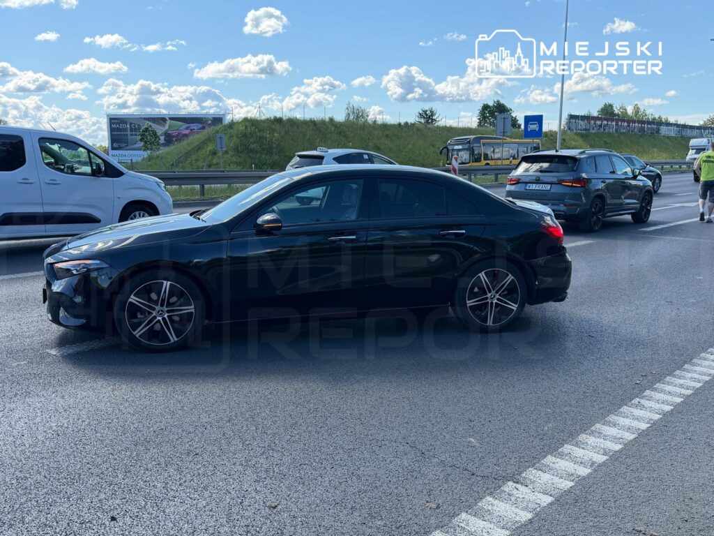 Czarny sedan zatrzymany na pasie drogowym wśród innych pojazdów, w tle billboard reklamowy.