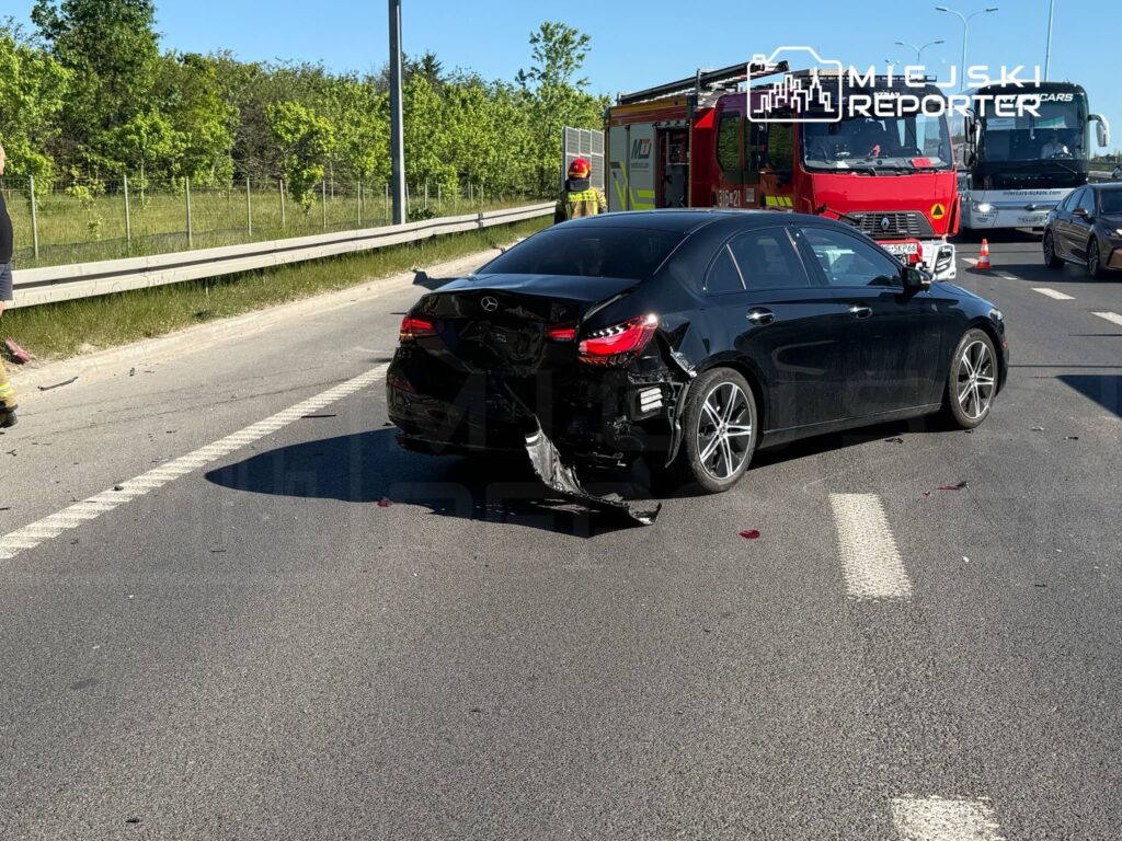 Czarny samochód osobowy z uszkodzoną tylną częścią stoi na jezdni, obok pracują strażacy i widać pojazd strażacki.