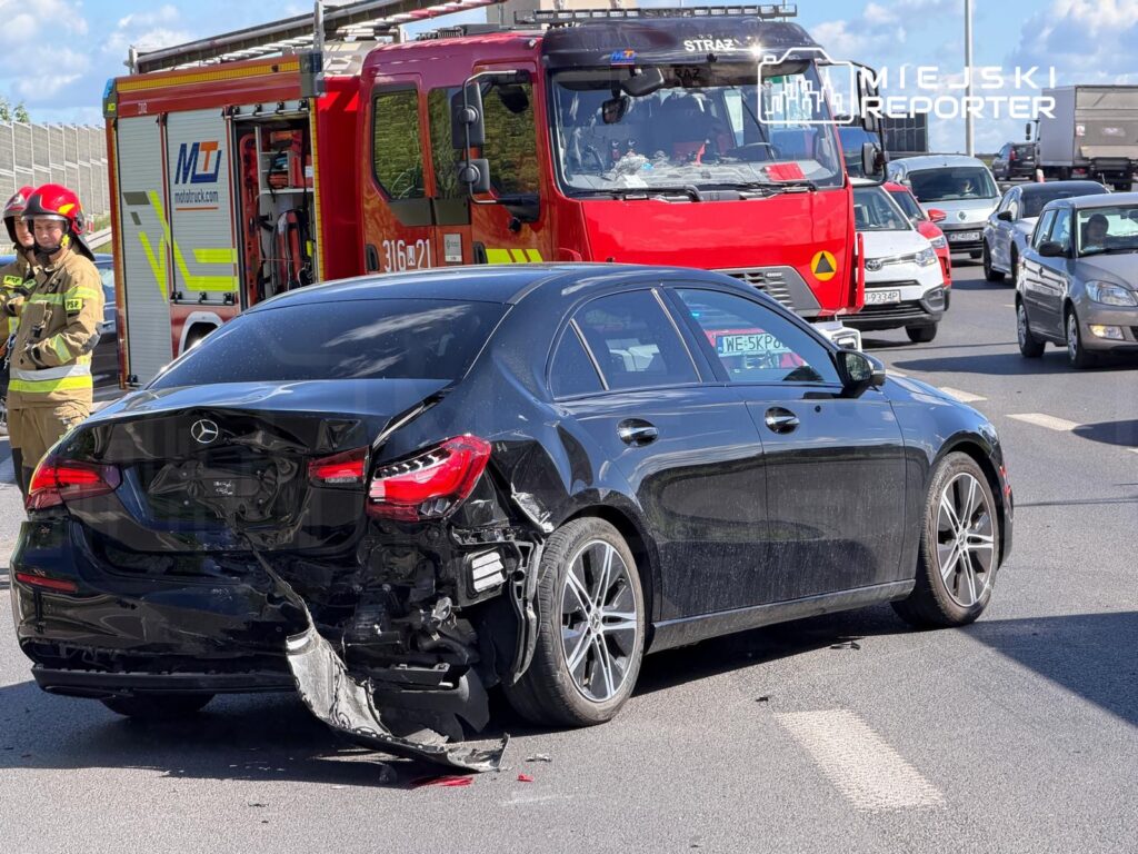 Czarny samochód osobowy z uszkodzeniami tyłu stoi na drodze, obok wozu strażackiego oraz strażaków w mundurach.