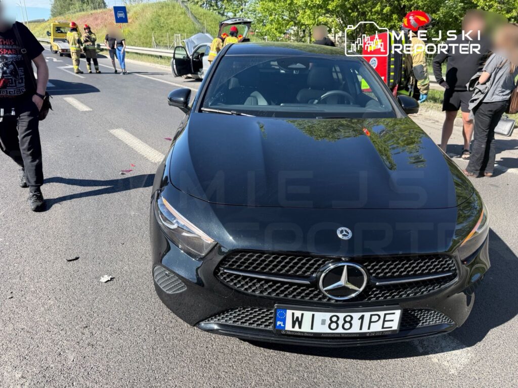 Czarny samochód osobowy marki Mercedes stoi na drodze, obok grupy osób oraz strażaków w mundurach.