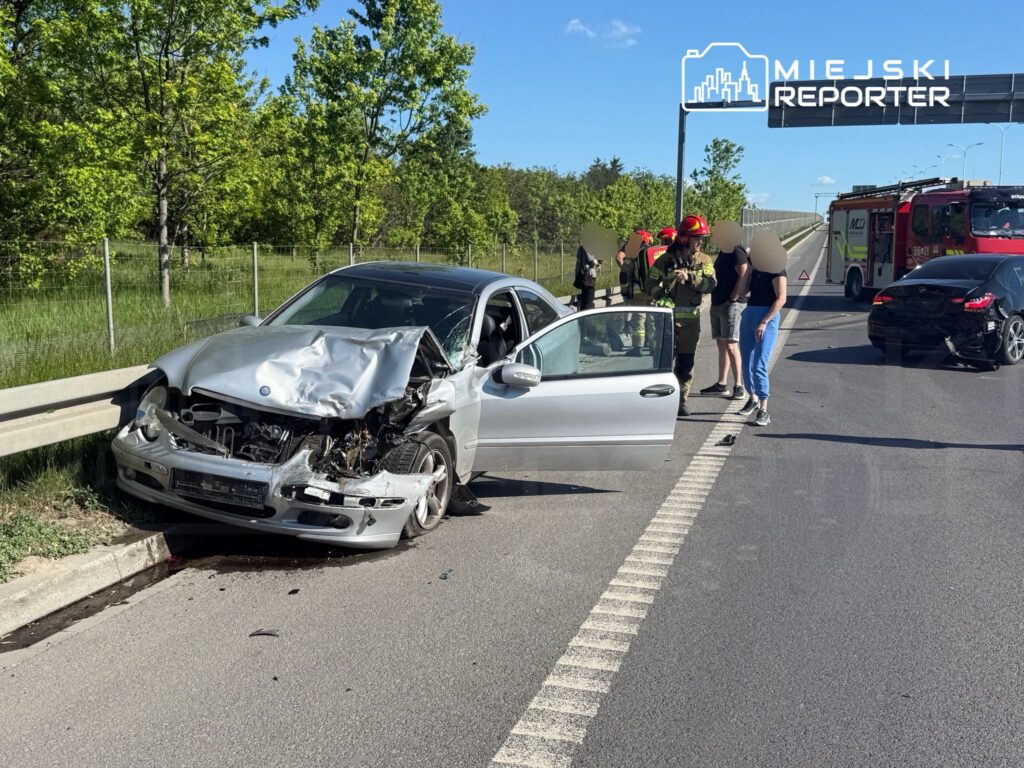 Uszkodzony samochód osobowy stoi przy drodze, obok grupy osób oraz strażaków w mundurach.