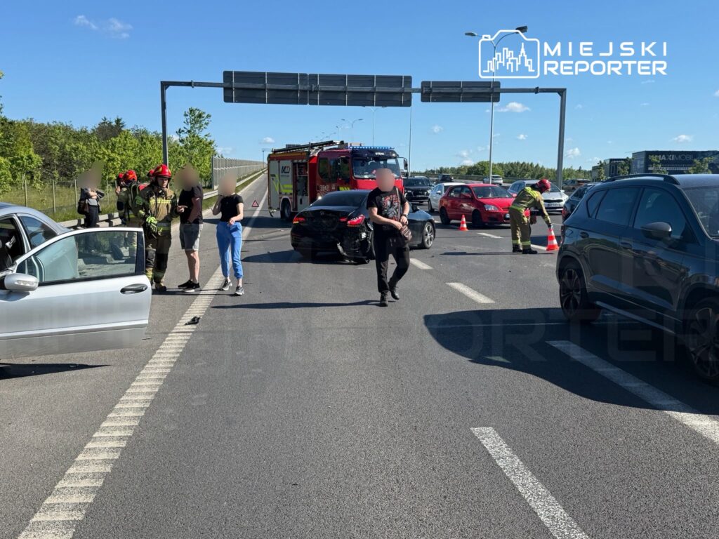 Na drodze doszło do wypadku, w którym uczestniczą dwa samochody; strażacy i osoby poszkodowane rozmawiają obok.