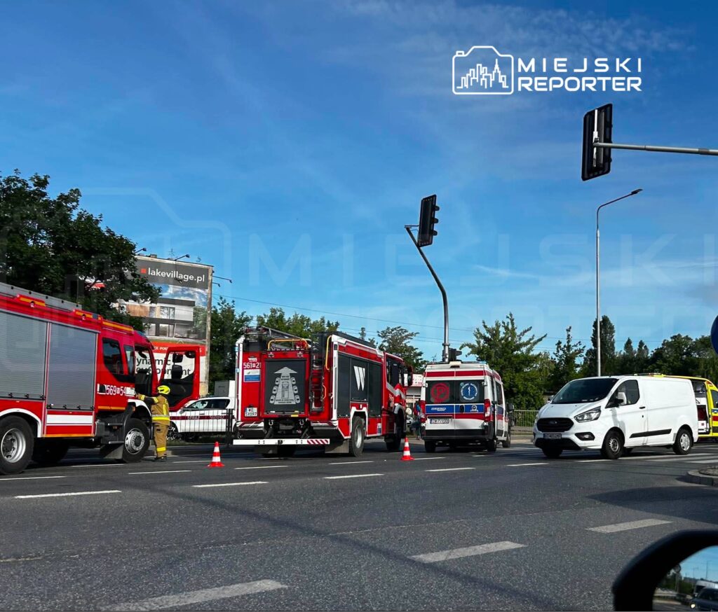 Na drodze stoją pojazdy straży pożarnej oraz ambulans, a w tle widoczna reklama z napisem "lakevillage.pl".