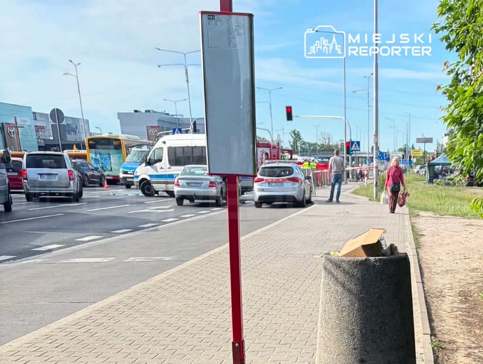 Ruchliwa ulica z samochodami stojącymi w korku, na pierwszym planie przystanek autobusowy oraz piesza osoba idąca w stronę chodnika.