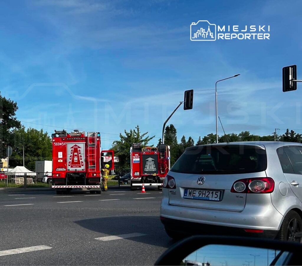 Czerwone wozy strażackie zatrzymane na skrzyżowaniu, a strażak kieruje ruchem w słoneczny dzień.