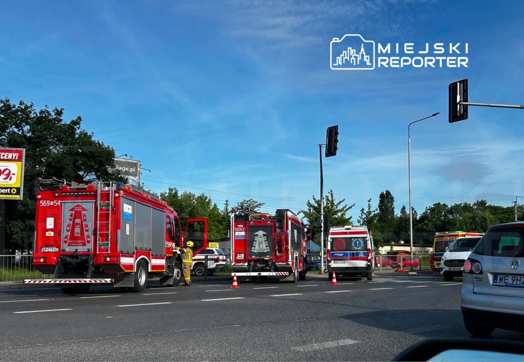 Czerwone wozy strażackie oraz ambulansy na ruchliwej ulicy, z ustawionymi pachołkami drogowymi.