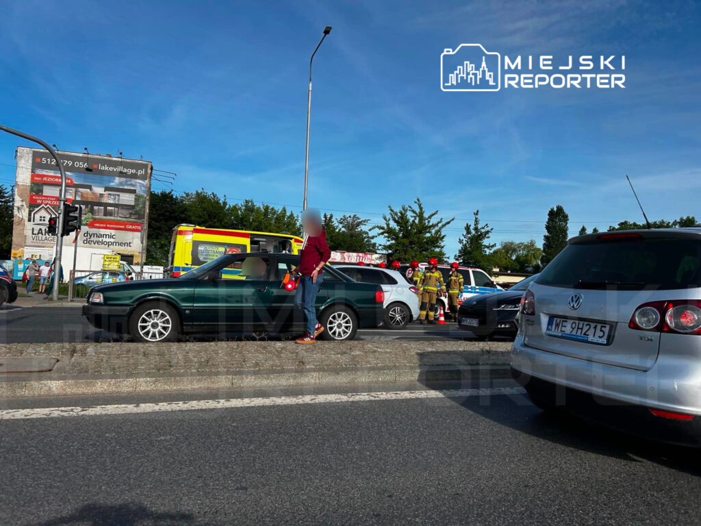 Kierowca zielonego samochodu stoi obok pojazdu na poboczu drogi, a w tle widać strażaków i karetki w trakcie akcji ratunkowej.