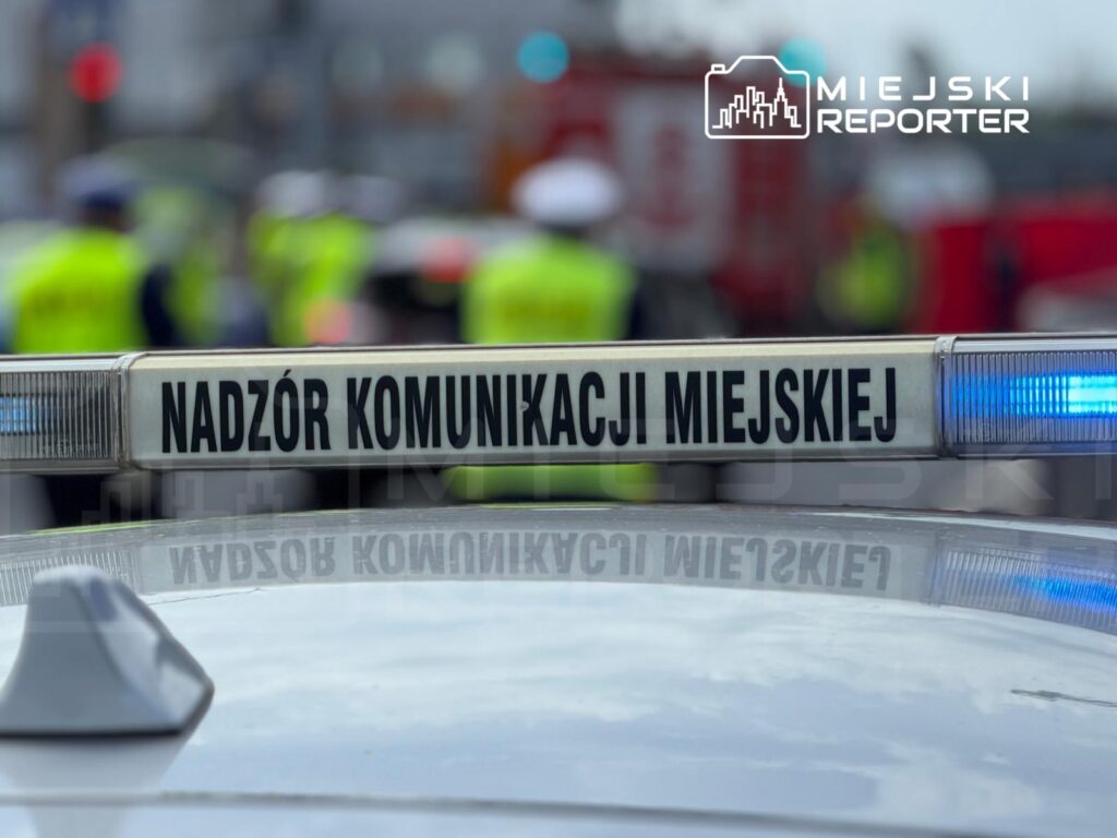 Na dachu radiowozu widoczny napis "NADZÓR KOMUNIKACJI MIEJSKIEJ" oraz niebieskie światło sygnalizacyjne, w tle funkcjonariusze w kamizelkach.
