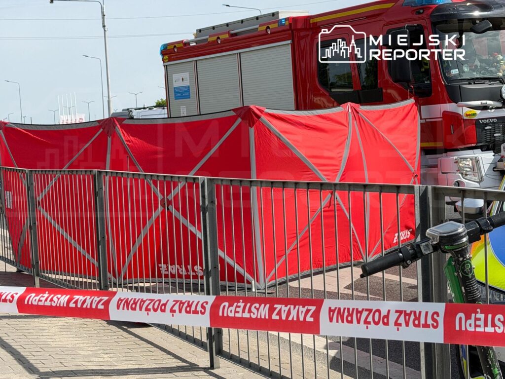 Czerwony namiot zabezpieczający ustawiony przy drodze, obok wozu strażackiego i roweru, z taśmą ostrzegawczą.