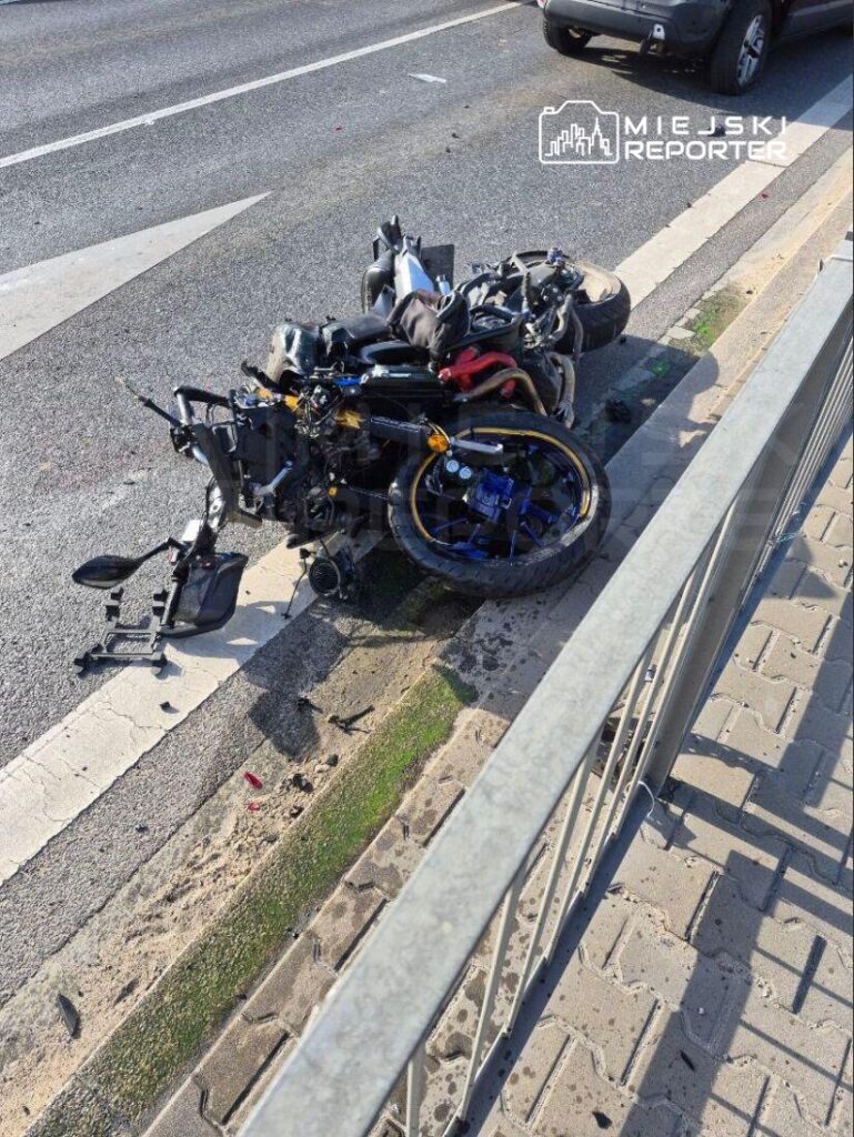 Zniszczony motocykl leży na poboczu drogi, obok metalowego ogrodzenia, z widocznymi śladami wypadku.