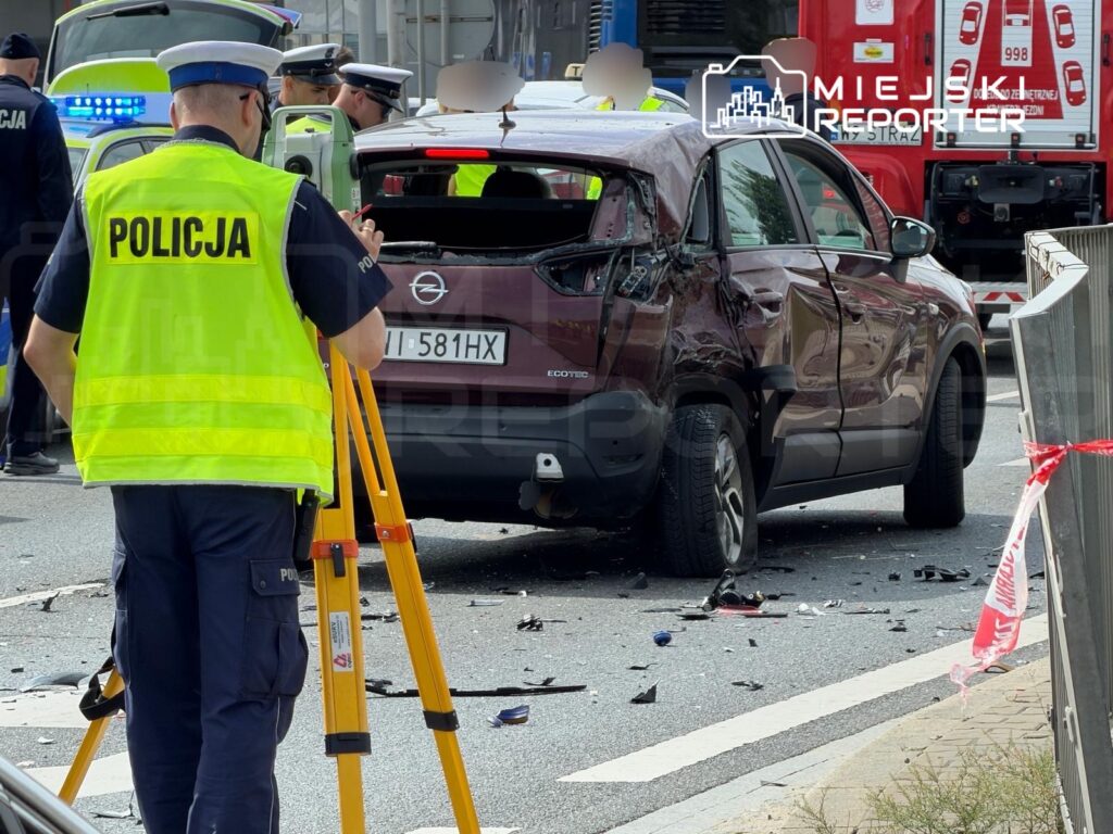 Policjant w żółtej kamizelce zabezpiecza miejsce wypadku drogowego z uszkodzonym samochodem osobowym.