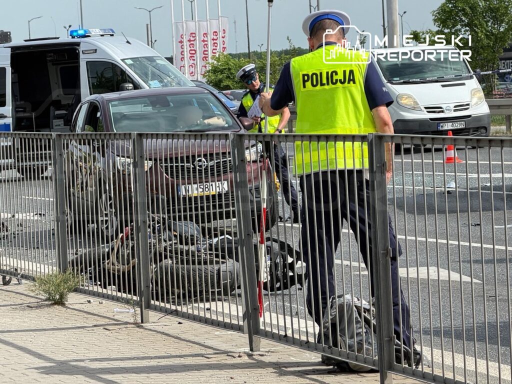 Policjanci badają miejsce wypadku drogowego obok uszkodzonego samochodu osobowego na ruchliwej ulicy.