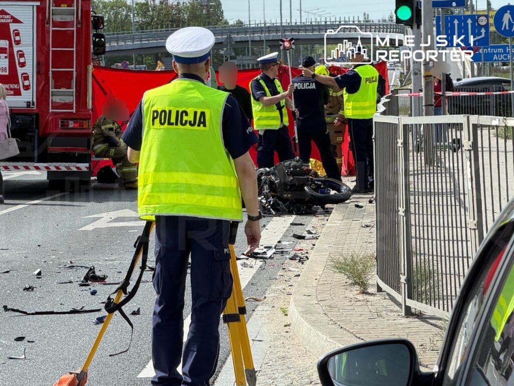 Policjanci i strażacy zabezpieczają miejsce wypadku drogowego, obok leży rozbity motocykl, na jezdni widoczne ślady zdarzenia.