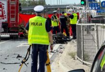 Policjanci i strażacy zabezpieczają miejsce wypadku drogowego, obok leży rozbity motocykl, na jezdni widoczne ślady zdarzenia.