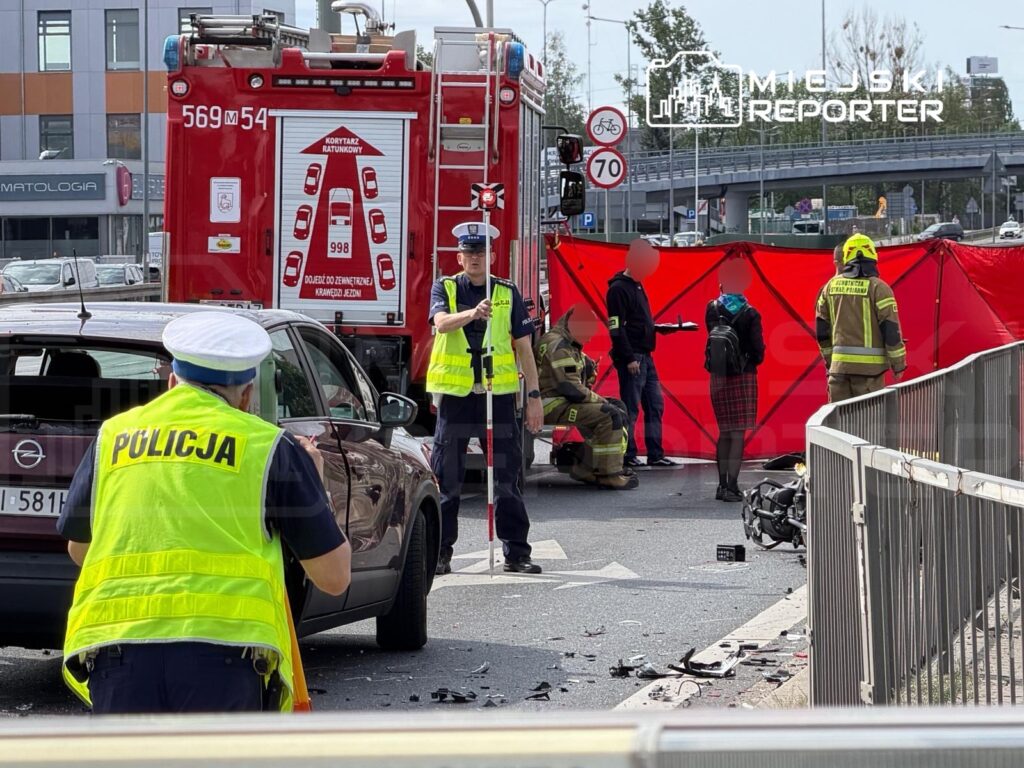 Policjant w odblaskowej kamizelce kieruje ruchem obok wypadku drogowego, z czerwonym namiotem zabezpieczającym miejsce zdarzenia.