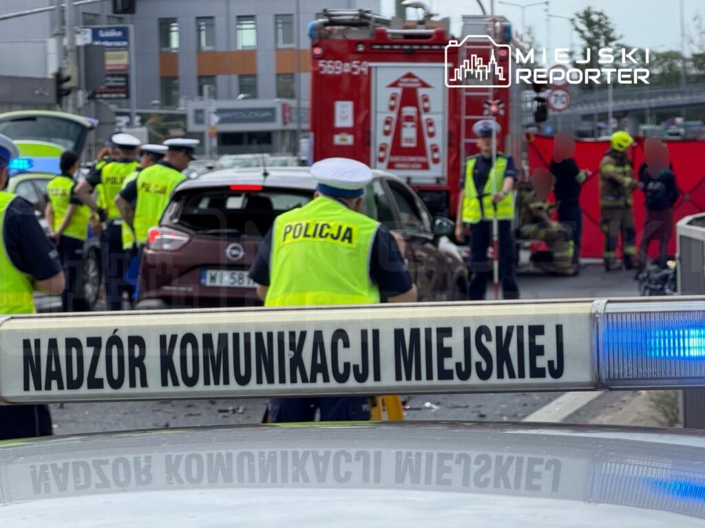 Grupa policjantów w kamizelkach odblaskowych zabezpiecza miejsce wypadku drogowego, w tle widoczne pojazdy i strażacy.