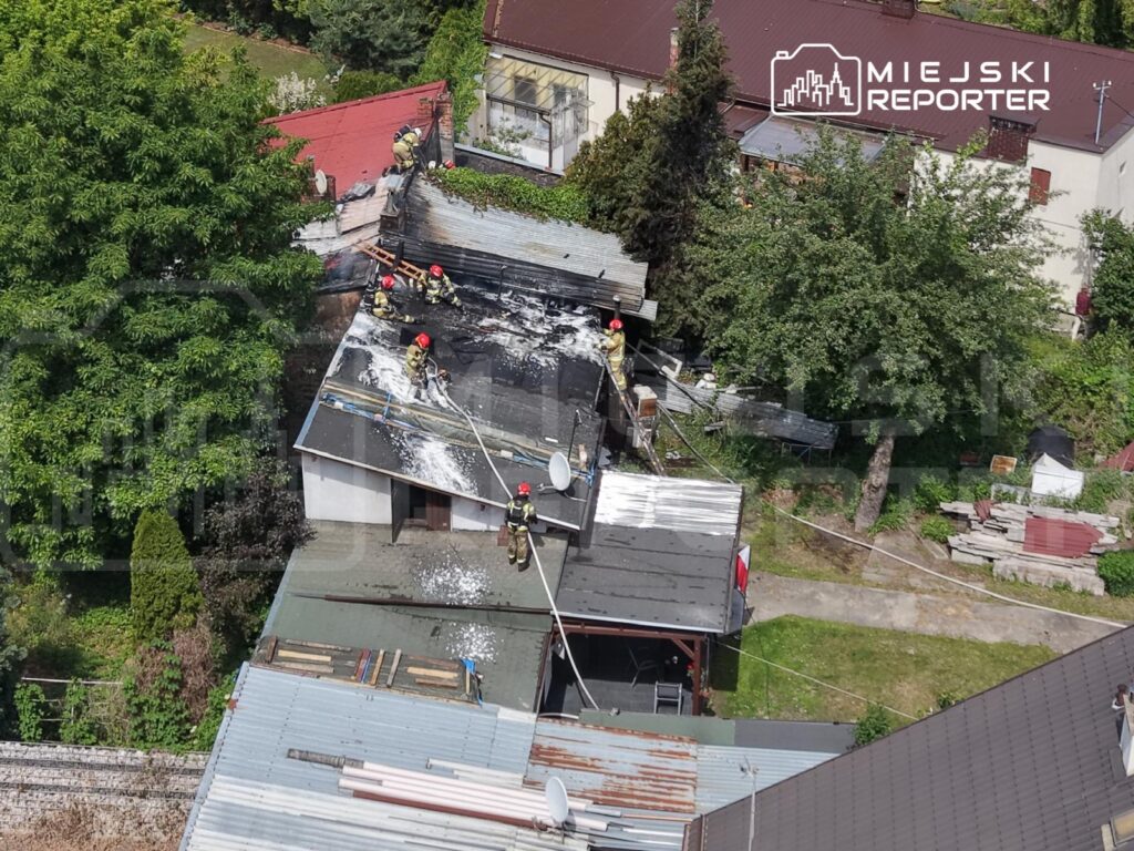 Grupa strażaków gasi pożar dachu na budynku mieszkalnym, otoczonym zielenią i innymi zabudowaniami.