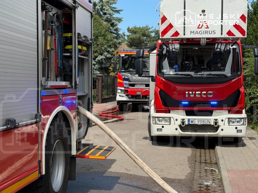 Wóz strażacki z rozwiniętym wężem pożarniczym na ulicy, obok drugi wóz strażacki w tle.