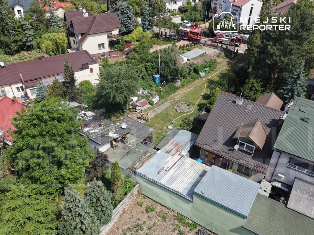 Grupa strażaków prowadzi prace na dachu budynku, w otoczeniu zieleni i sąsiednich domów. W tle widoczne wozy strażackie.