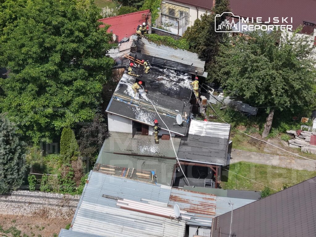 Grupa strażaków gasi pożar dachu budynku mieszkalnego, używając węża strażackiego i sprzętu ochronnego.