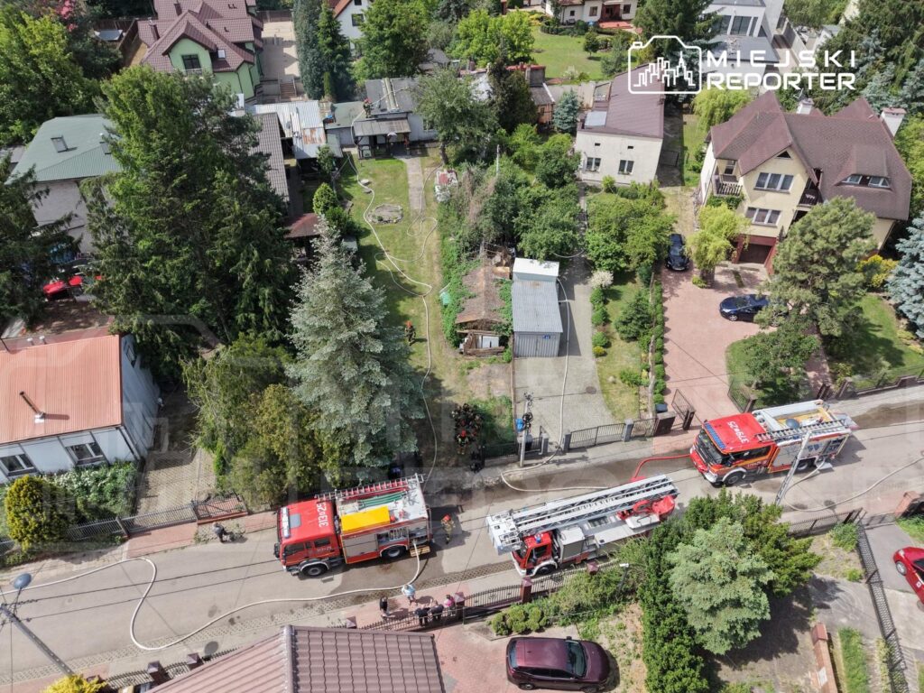 Dwa wozu strażackie stoją na ulicy, a strażacy rozkładają węże w otoczeniu domów jednorodzinnych i zieleni.