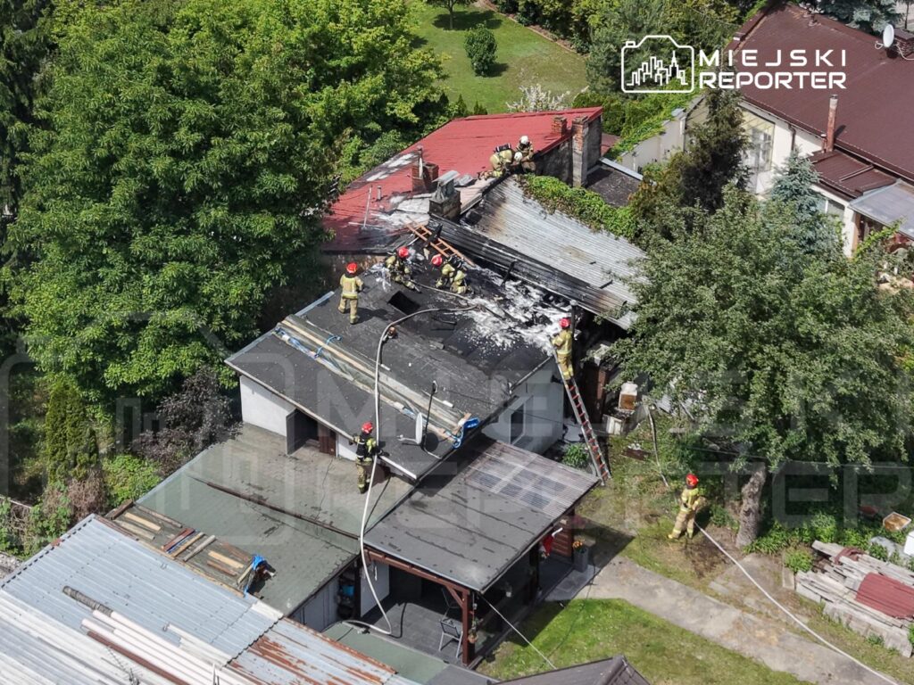 Strażacy gaszą pożar na dachu budynku, używając węża strażackiego, w otoczeniu zieleni i sąsiednich domów.