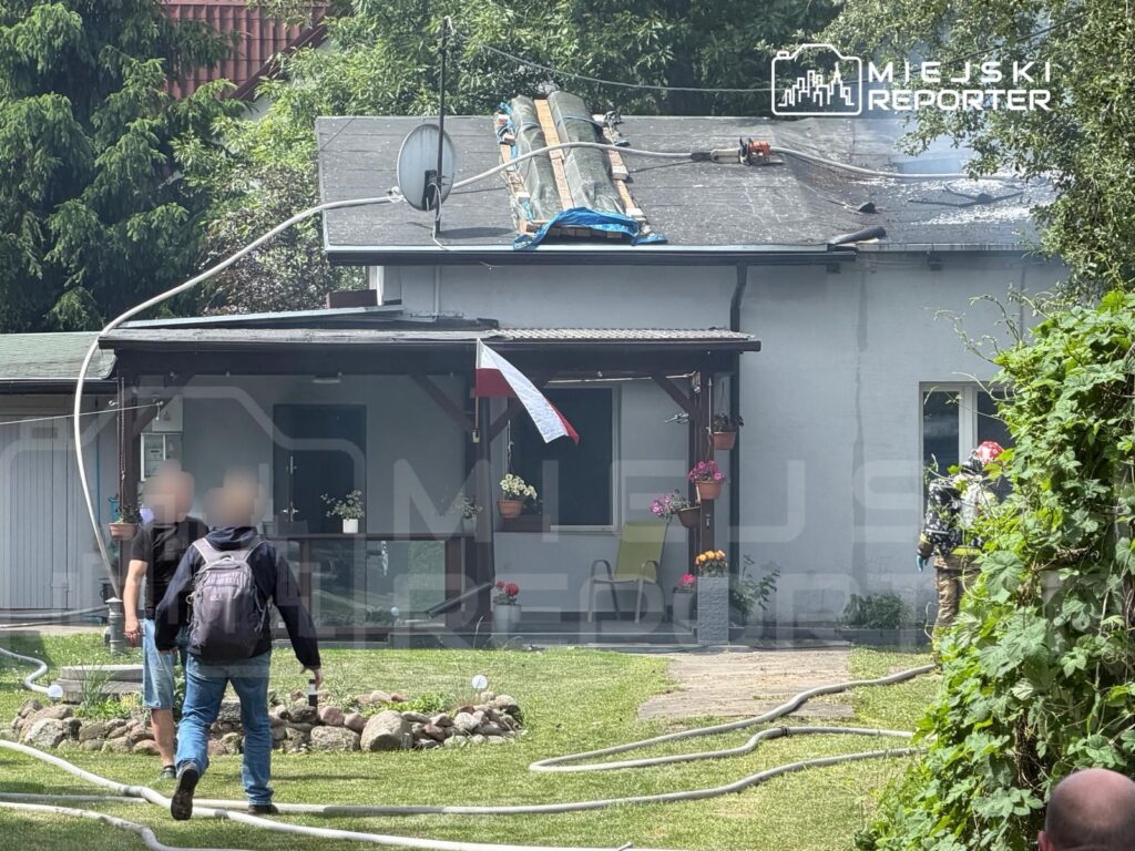 Dwaj mężczyźni idą w kierunku budynku z dymiącym dachem, obok leżą węże strażackie na trawniku.