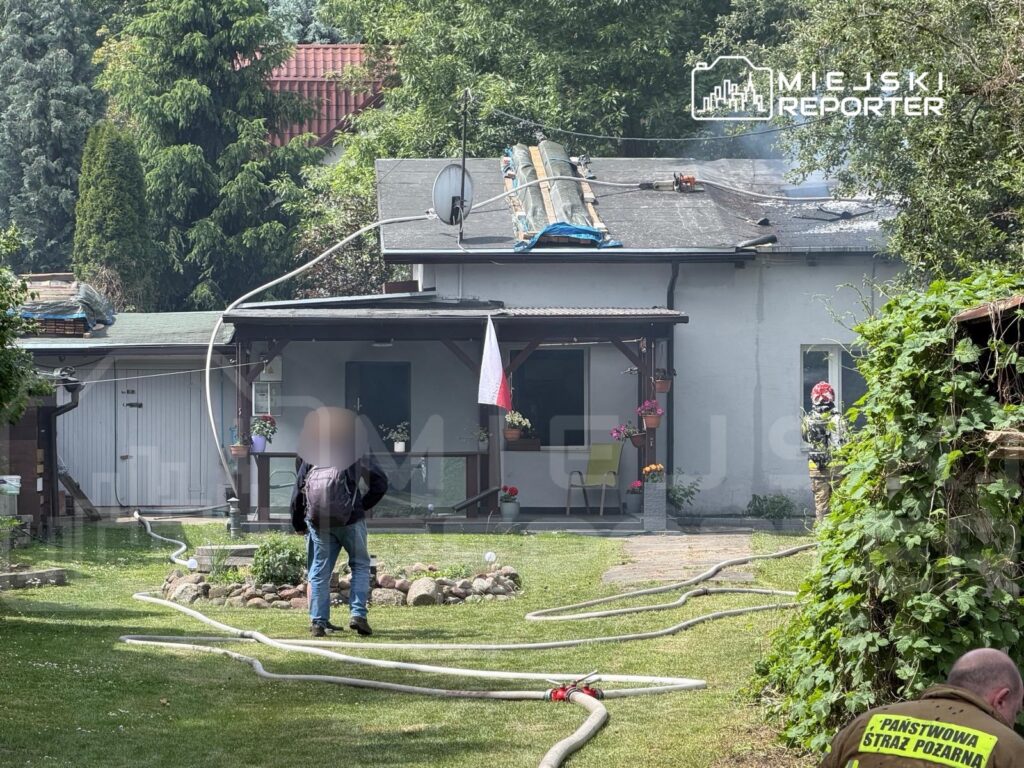Mężczyzna w czarnej kurtce obserwuje działania strażaków przy budynku, z którego wydobywa się dym.
