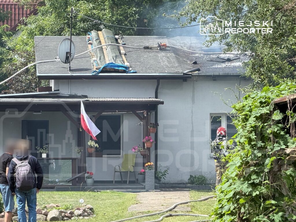 Dym unoszący się z dachu domu jednorodzinnego, obok grupka ludzi obserwujących sytuację.