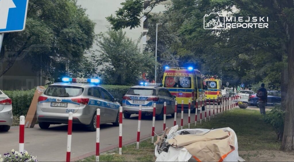 Na chodniku parkują radiowozy policyjne oraz karetki pogotowia z włączonymi sygnałami świetlnymi.