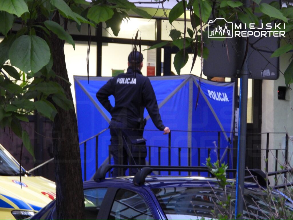 Policjant w mundurze stoi przy niebieskim namiocie z napisem "POLICJA" w miejskim otoczeniu.