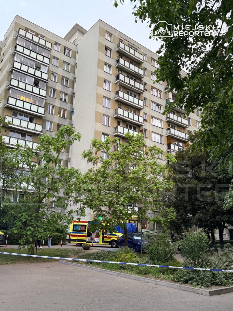 Wielopiętrowy budynek mieszkalny z balkonami, w tle karetka pogotowia i policja na miejscu zdarzenia.