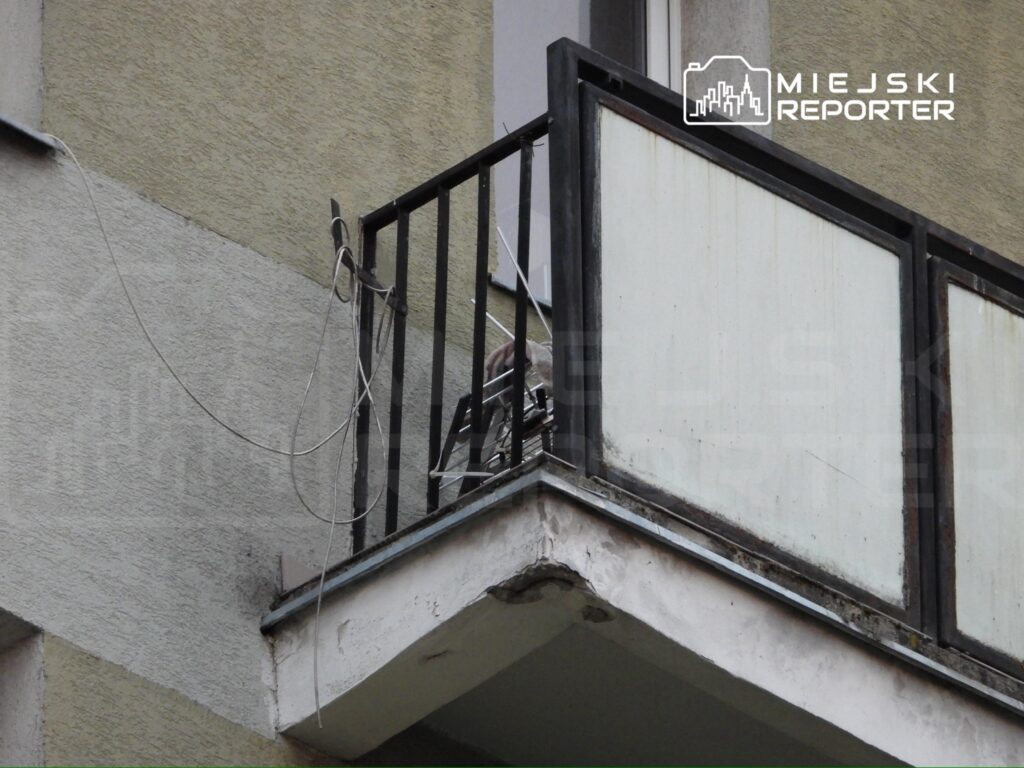 Balkon z metalowym ogrodzeniem, na którym widoczne są luźne kable oraz zniszczone panele.