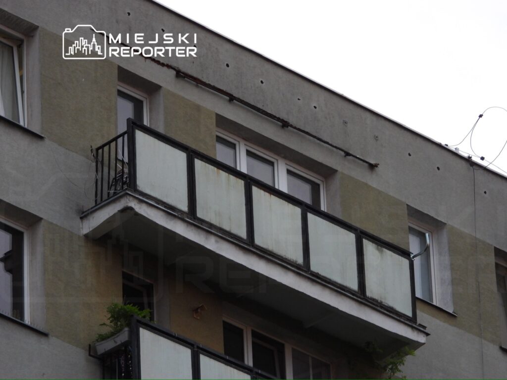 Balkon mieszkania z metalowym balustradą, na którym stoi krzesło i rośnie roślina, przy szarej elewacji budynku.
