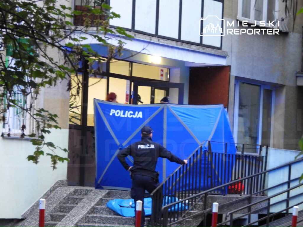 Policjant przy niebieskim namiocie zabezpieczającym miejsce zdarzenia przed wejściem do budynku.