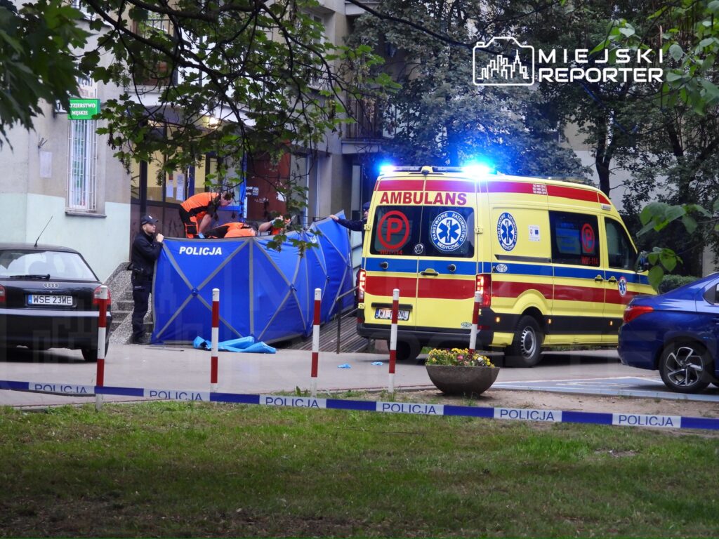 Grupa ratowników medycznych w pomarańczowych uniformach udziela pomocy przy wejściu do budynku, obok parkujący ambulans.
