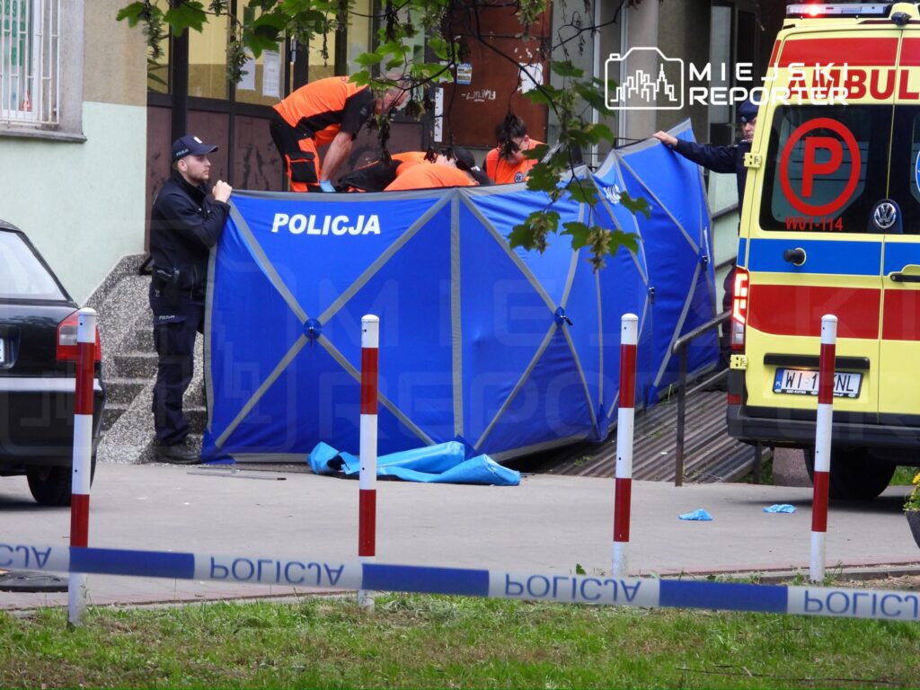 Policjant nadzorujący prace ratowników w pomarańczowych strojach, ustawiających niebieski namiot na chodniku.